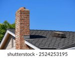 A residential roof with a red brick chimney with its chimney cap lying displaced on the black shingles.