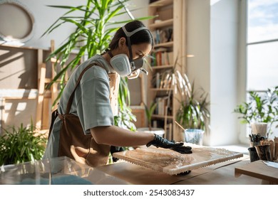 Resin art. Woman artist applying epoxy resin with gloved fingers in art studio workshop. Focused female wearing protective gloves removes excess uncured resin from painting with fingers. Fluid art. - Powered by Shutterstock