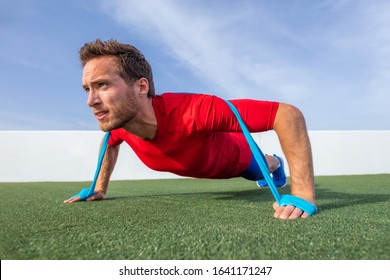 Resistance bands fit man doing pushups with elastic band as extra difficulty at outdoor gym park. Athlete doing dynamic workout pulling rubber elastics plyometrics exercises.