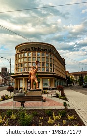 Resita, Romania-May 25, 2021: The Old Building Of The Universal Store With Drawn Windows And The Statue Of The Worker In Front Of It, Photographed When Two People Were Sitting Quietly On The Bench