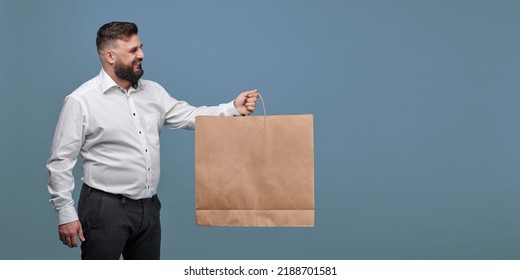 A Respectable Man Holds Out A Brown Paper Bag. A Man In A White Shirt With A Paper Bag On A Blue Background Looks To The Side. The Man Looks At The Paper Bag. Advertising Studio Photo