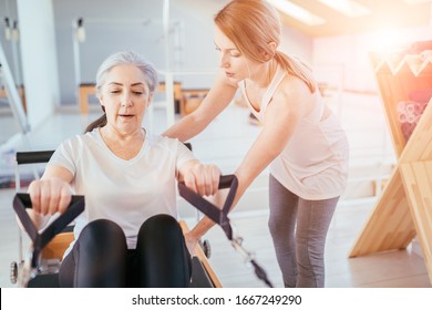 Respectable old age grey hair woman workout pilates with personal trainer on reformer practice in pilates studio, working out indoor, correcting beginners, rehabilitation concept.n Sun glare effect.