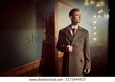 Respectable young man in an elegant coat stands in a vintage style apartment with luxury interior. Luxury lifestyle. Men's beauty, fashion. Copy space.
