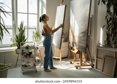 Responsible female artist compares two canvases organizes workspace puts things in order at workplace. Woman painter prepares for work day, performing actions necessary for productive creative process - Powered by Shutterstock