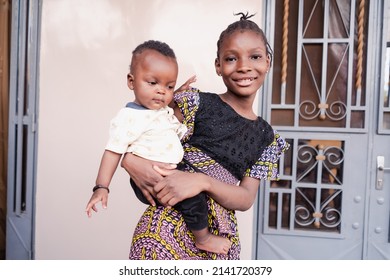 Responsible proud big sister takes care of her little brother, whom she carries in her arms as a matter of course according to African tradition and family ties - Shutterstock ID 2141720379