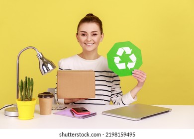 Responsible Self Confident Woman Holding Green Recycling Sign In Hand And Cardboard Package, Sitting On Workplace With Laptop. Indoor Studio Studio Shot Isolated On Yellow Background.