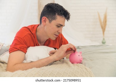 Responsible young caucasian dad worried about economic situation, laying down on his bed and putting a coin into a pink piggybank. Symbolic shot. High quality photo