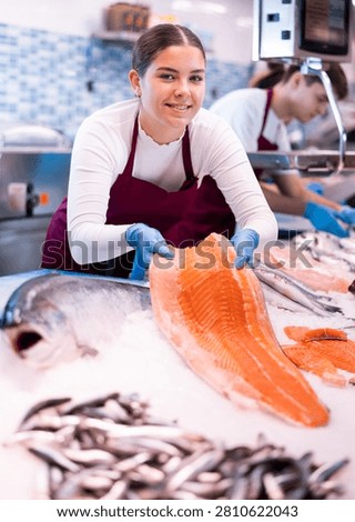 Responsive shop assistant keeping in hands fresh salmon in fish store with large assortment
