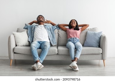 Rest Concept. Happy African American couple sitting on comfortable couch at home in living room. Cheerful casual guy and lady relaxing on sofa, leaning back, enjoying weekend free time, taking break - Powered by Shutterstock