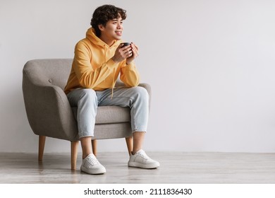 Rest concept. Happy Asian guy sitting in comfortable armchair and drinking coffee against white studio wall, free space. Cheerful casual man relaxing, enjoying free time or break with hot beverage - Powered by Shutterstock