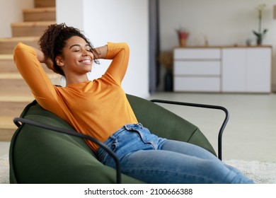 Rest Concept. Happy black lady sitting on comfortable bean bag at home in living room. Cheerful casual woman with closed eyes relaxing on sofa, leaning back, enjoy weekend free time or break from work