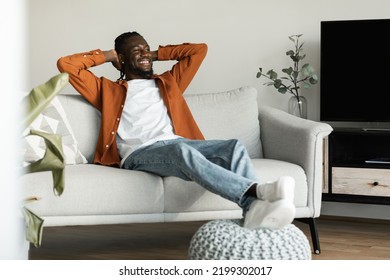 Rest concept. Happy black man sitting on comfortable couch at home in living room, leaning back with hands behind head, enjoying weekend free time or break from work - Shutterstock ID 2199302017