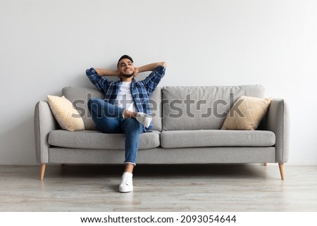 Rest Concept. Happy Middle Eastern guy sitting on comfortable couch at home in living room. Cheerful casual man relaxing on sofa, leaning back, enjoying weekend free time or break from work