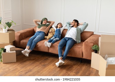 Rest Concept. Happy muslim family leaning back on the sofa in new house, open cardboard boxes on the floor. Happy dad, mum in hijab and little daughter lying on couch and looking at each other