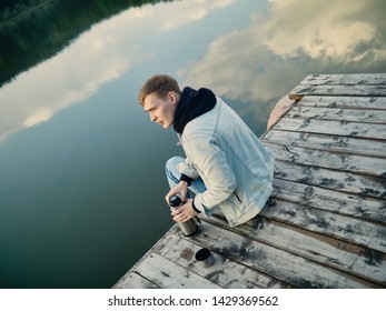 Rest on the lake. A young guy in blue jeans, a denim jacket and a black hoodie is sitting on a wooden pier against the background of the lake and the forest with a thermos in his hands.