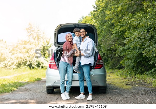 Rest outdoor from road, family journey at\
summer. Satisfied millennial arab man and lady in hijab holding\
baby near car with open trunk, copy space. Trip, travel together,\
vacation and people\
emotions