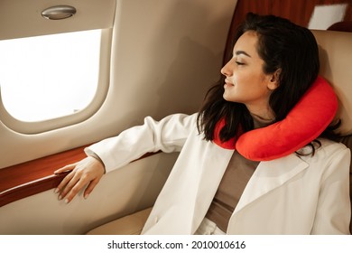 Rest In The Plane. A Young Girl In A White Suit Is Resting And Looking In The Window Of A Business Plane, Using A Red Neck Pillow For Travel