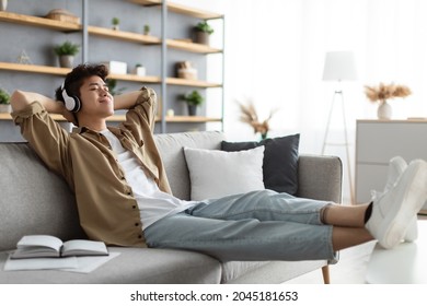 Rest And Relax Concept. Calm asian man sitting on couch, listening to music, audio book, podcast, enjoying meditation for sleep and peaceful mind in wireless headphones, leaning back, copy space