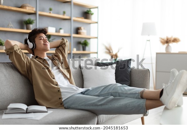 Rest And Relax Concept. Calm asian man sitting on\
couch, listening to music, audio book, podcast, enjoying meditation\
for sleep and peaceful mind in wireless headphones, leaning back,\
copy space