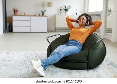 Rest And Relax Concept. Calm black woman sitting on bean bag, listening to music, audio book, podcast, enjoying meditation for sleep and peaceful mind in wireless headphones, leaning back, copy space