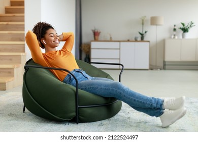 Rest And Relax Concept. Calm black woman sitting on bean bag, listening to music, audio book, podcast, enjoying meditation for sleep and peaceful mind in wireless earphones, leaning back, copy space - Powered by Shutterstock