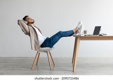 Rest And Relax Concept. Happy Arab man sitting on chair, listening to music, audio book, podcast in wireless headphones, leaning back sitting at desk with legs on table and hands behind head