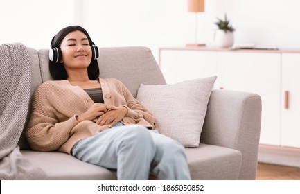 Rest And Relax Concept. Portrait Of Calm Asian Woman Lying On The Couch, Listening To Music, Audio Book Or Podcast, Enjoying Meditation For Sleep And Peaceful Mind In Wireless Headset At Home