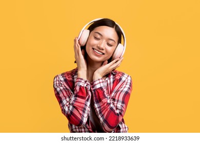 Rest And Relax Concept. Positive Chinese Woman Wearing And Touching Wireless Headset, Listening To Music With Closed Eyes Over Yellow Studio Background, Copy Space