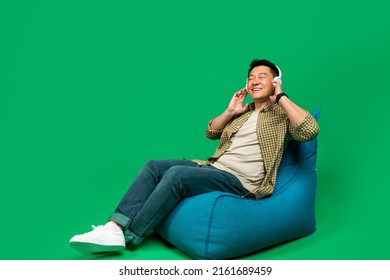 Rest And Relax. Mature Asian Man Sitting On Beanbag Chair And Listening To Music With Closed Eyes, Enjoying Great Sound In New Wireless Headphones, Green Background, Free Space