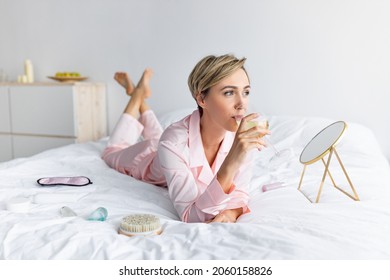 Rest And Relax. Portrait Of Middle-aged Lady Lying In Bed On White Bedsheets Holding Glass Of Alcohol Beverage Drinking Wine Or Champagne And Looking Away. Celebration At Home Alone