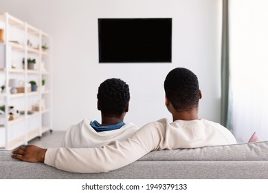 Rest And Relax. Rear Back View Of Afro Man And Woman Sitting On The Sofa In Living Room, Watching Television Show Together, Looking At Blank Plasma Tv On The Wall With Isolated Empty Monitor, Mock Up