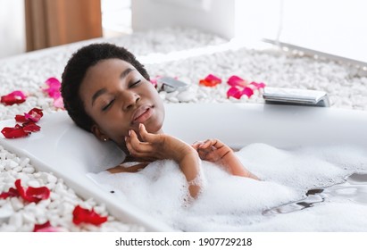 Rest, relaxation and beauty day at weekend or vacation in morning. Happy millennial african american woman lies in bathtub with flower petals in white bathroom interior in daylight, empty space