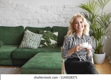 Rest And Relaxation Concept. Portrait Of Attractive Successful 55 Year Old Female CEO Having Coffee Break, Holding Mug And Plate, Smiling Broadly At Camera, Sitting On Chair In Modern Office Interior