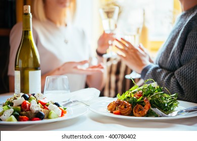 Restaurant Or Cafe Table With Plate Of Salads And Wine. Two People Talking On Background