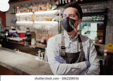 Restaurant Employee Wearing A Protective Mask Against The Covid Pandemic Virus Cafe Bistro. The Waiter Is A Young Man Of Dark Caucasian Appearance Standing Near The Bar. Workman's Clothing In Apron An