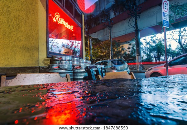 Restaurant facade,\
bright red sign with beer barrels on the sidewalk in a rainstorm.\
Wet corner bar\
table.