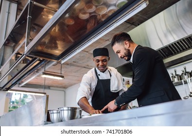 Restaurant manager discussing with chef in kitchen. Cook preparing a dish with restaurant owner standing by.