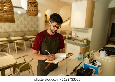 Restaurant manager jotting down notes in a notebook while standing at the counter, focusing on daily tasks and planning for the business