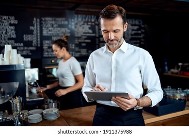 Restaurant Manager Standing With Digital Tablet With Employee In Background Preparing Coffee