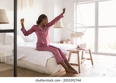 Rested African American Woman Yawning And Stretching Hands After Good Sleep Sitting On Bed In Bedroom, Wearing Pajamas. Happy Well-Slept Female Waking Up Enjoying Weekend Morning At Home