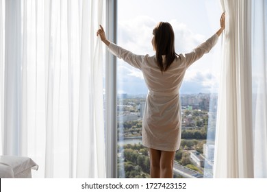 Rested female in white night robe wake up opens curtains enjoys big city and sunny weather, rear view. Welcoming of new day, good morning, modern warm hotel bedroom, luxury apartments owner concept