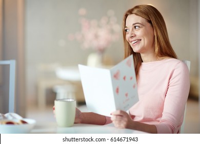 Restful young woman with cup of drink reading greeting-card