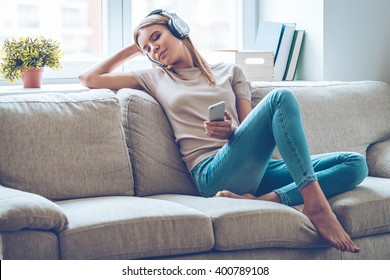 Resting With Joy. Beautiful Young Woman Listening To Music And Keeping Eyes Closed While Sitting On Sofa At Home