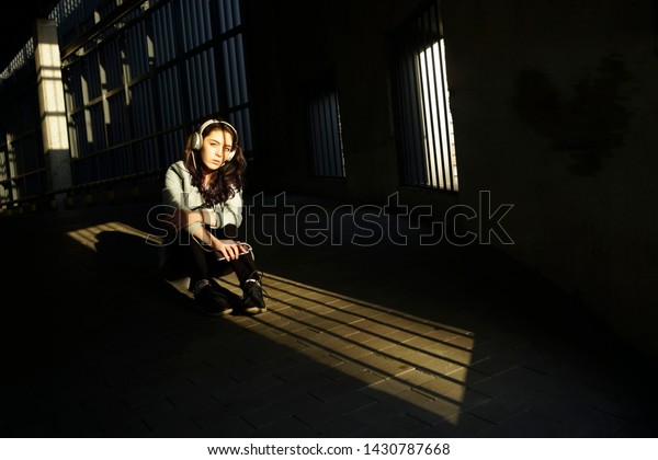 Resting with joy. Beautiful young woman listening to\
music and sitting on floor at car parking.sunny light and shadows\
on face