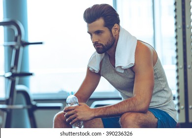 Resting Time. Handsome Young Men In Sports Wear Wearing Towel On His Shoulders And Holding Water Bottle While Sitting At Gym