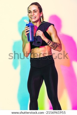 Resting time. Sporty girl with towel on shoulder drinks water. Photo of muscular woman in black sportswear on white background with effect of rgb colors shadows. Sports and healthy lifestyle
