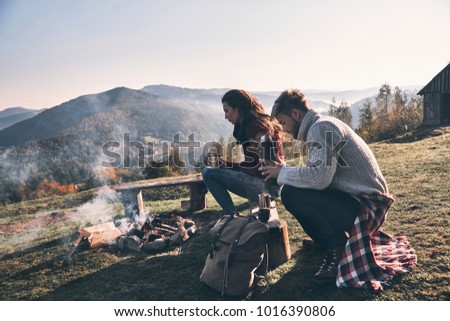 Resting together. Beautiful young couple having morning coffee while sitting by the campfire in mountains