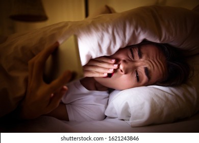 A Restless Sleeper. Close-up Photo Of A Girl, Who Is Yawning While Checking Notifications On Her Smartphone, Lying In Her Bed Covered With A Blanket Up To Her Head.