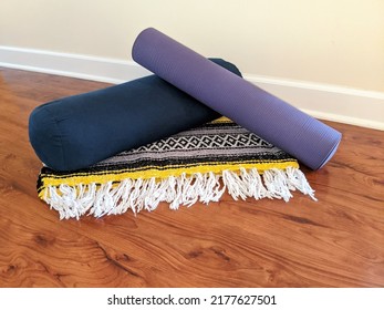 Restorative yoga props on a wooden laminate floor with a neutral wall background. Props include a purple yoga mat, a navy bolster, and a patterned blanket.