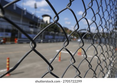 Restricted background view of an empty racetrack and grandstand seen through metal chain link fence under blue sky, creating feeling of anticipation and confinement